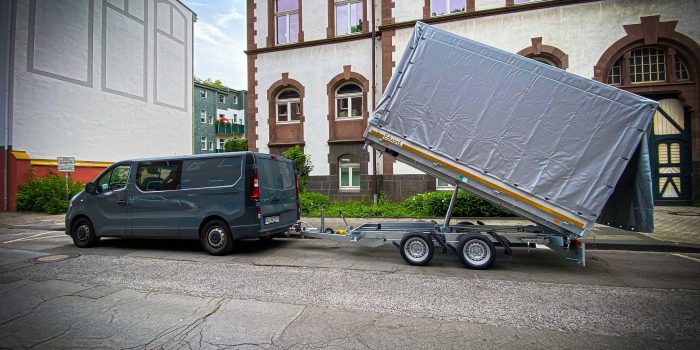 Ein grauer Lieferwagen, der einen Anhänger mit einer abgedeckten Ladung zieht, steht auf einer Straße vor einem Backsteingebäude mit gewölbten Fenstern und Türen