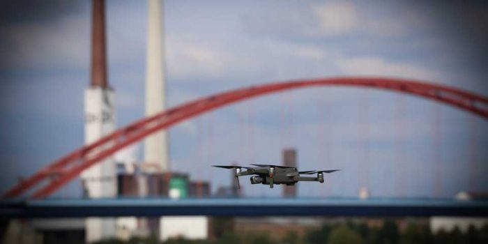 Eine Drohne fliegt durch die Luft, im Hintergrund sind eine rote Bogenbrücke und eine Stadtlandschaft zu sehen.